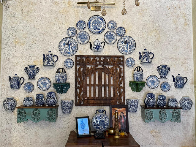 Wall display of blue and white ceramic items including plates, vases, and teapots against a textured wall.